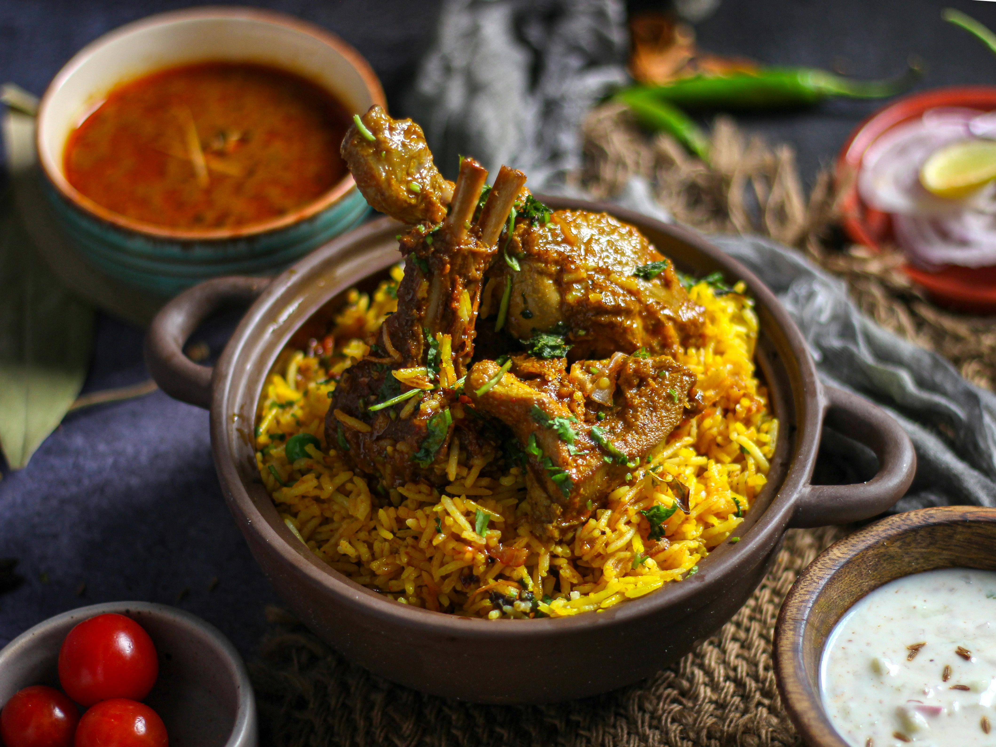 Image of biryani in bowl with chicken leg pieces arranged on top with raitha and sheerwa beside the bowl.